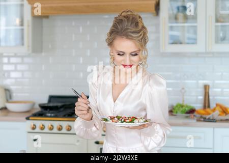 Kaukasische schöne Frau bereitet frischen Salat reife Erdbeeren und Rucola Blätter, Diät-Frühstück luxuriöse Mädchen in einem schönen hellen Kleid mit Make-up und Frisur. Glücklich in der Küche zu Hause Stockfoto