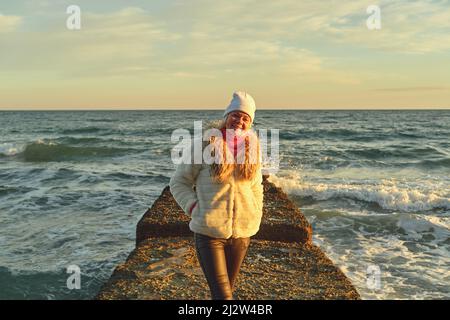 Eine Frau mit blondem Haar in einem Hut und einem Pelzmantel geht an einem Steinbrecher entlang des Meeres Stockfoto