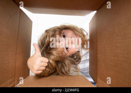 Überrascht Kind Junge mit Daumen nach oben Auspacken, Öffnen Karton und Blick nach innen. Das Paket, Lieferung, Überraschung, Geschenk für Kind. Emotionen von Kindern Stockfoto