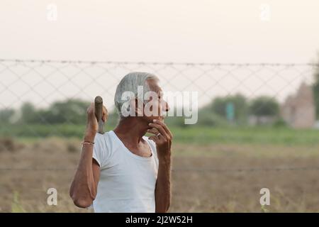 Nahaufnahme eines älteren indischen Bauern, der an etwas denkt, während er bei Sonnenuntergang eine Schaufel in der Hand hält Stockfoto