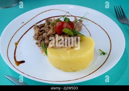 Fleischbällchen mit Kartoffelpüree auf einem weißen Teller. Fleisch in saurer Rahmsoße. Tomaten und Basilikum. Essen im Restaurant. Mittagspause. Medaillons. Stockfoto