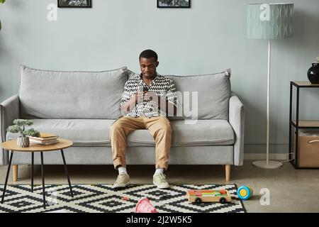 In voller Länge Porträt eines jungen schwarzen Mannes mit Smrartphone beim Entspannen auf der Couch allein zu Hause in minimalem grauen Innenraum, Kopierraum Stockfoto