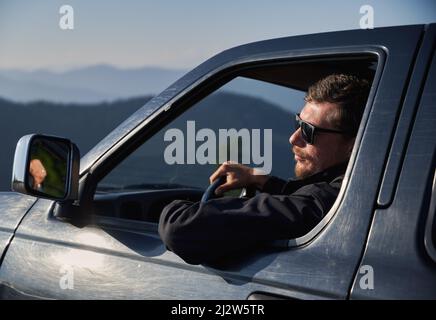 Nahaufnahme eines Mannes in einer Sonnenbrille, der ein schwarzes Auto fährt und nach links auf die Berglandschaft blickt, während er am Nachmittag in den Bergen bei Sonnenschein fährt. Stockfoto