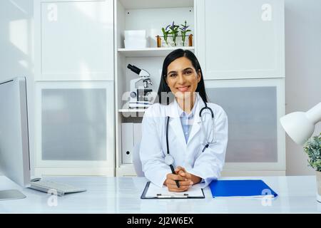 Porträt einer schönen jungen Ärztin in weißer Laborkittel-Uniform, die am Schreibtisch in ihrer modernen Büro-Gesundheitsbranche sitzt Stockfoto