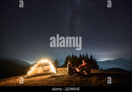 Frau mit Gitarre in den Händen, die am Lagerfeuer neben ihrem lichtdurchfluteten Zelt gegenüber dem Mann sitzt. Romantischer Abend mit Liebsten im Freien unter Sternenhimmel in den Bergen. Konzept des Nachtcampens. Stockfoto