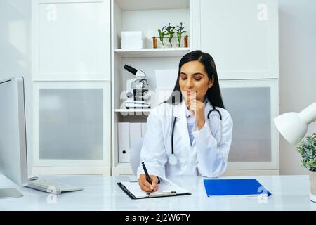 Porträt einer schönen jungen Ärztin in weißer Laborkittel Uniform, die in ihrer modernen Bürobranche im Gesundheitswesen am Schreibtisch arbeitet Stockfoto