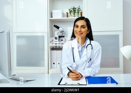 Porträt einer schönen jungen Ärztin in weißer Laborkittel-Uniform, die am Schreibtisch in ihrer modernen Büro-Gesundheitsbranche sitzt Stockfoto