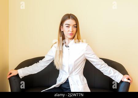Porträt eines jungen kaukasischen Arztes mit blondem, langen Haar und weißem Mantel, der auf dem schwarzen Sofa sitzt und die Kamera anschaut Stockfoto