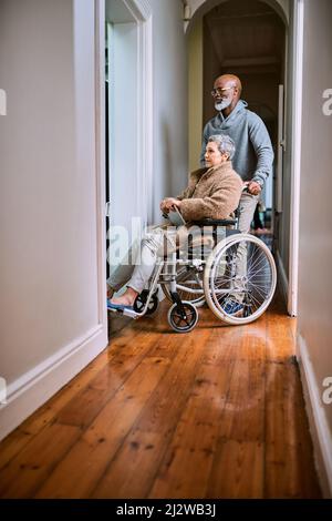 Hilft ihr dabei, sich zu bewegen. Ausgeschnittene Aufnahme eines älteren Mannes, der seine Frau mit Rollstuhl durch das Haus schiebt. Stockfoto