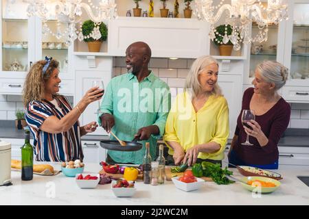 Glückliche kaukasische ältere Frauen, die Wein trinken, während Freunde zu Hause gemeinsam Essen zubereiten Stockfoto