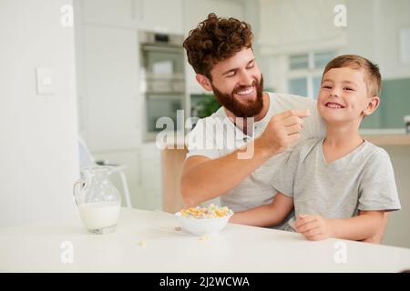 Das Frühstück bringt das erste Lächeln des Tages. Aufnahme eines entzückenden kleinen Jungen und seines Vaters beim gemeinsamen Frühstück zu Hause. Stockfoto