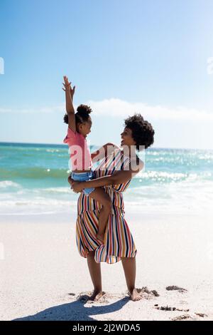 Fröhliche afroamerikanische Mutter, die ihre Tochter trägt, während sie den sonnigen Tag am Strand gegen den Himmel genießt Stockfoto