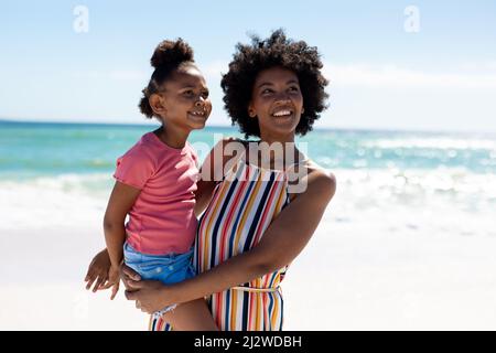 Glückliche afroamerikanische Mutter, die Tochter trägt, während sie das Sommerwochenende am Strand gegen den Himmel genießt Stockfoto