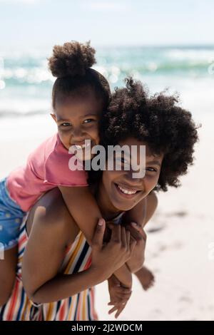 Porträt einer glücklichen afroamerikanischen Frau, die an sonnigen Tagen am Strand der Tochter Huckepack gibt Stockfoto