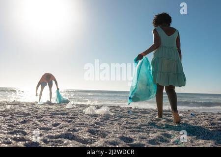 Die ganze Länge des afroamerikanischen Paares pflückt Abfall auf Sand am Strand gegen klaren blauen Himmel Stockfoto