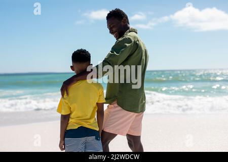 Rückansicht eines glücklichen afroamerikanischen Vaters und Sohnes, der an einem sonnigen Tag am Strand spazieren ging Stockfoto