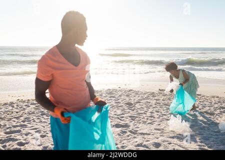 afroamerikanischer Mann hält Plastiktüte, während er seine Freundin anschaut, die Müll am Strand sammelt Stockfoto