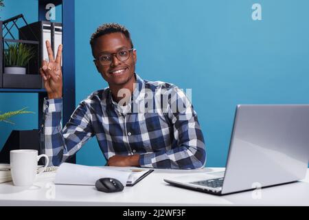 Lächelnder, fröhlicher Mann, der im Büro mit der Hand das Siegersymbol macht. Fröhliche, fröhliche afroamerikanische Büroangestellte, die mit den Fingern eine Siegesfriedensgeste macht, während sie die Kamera anschaut. Stockfoto