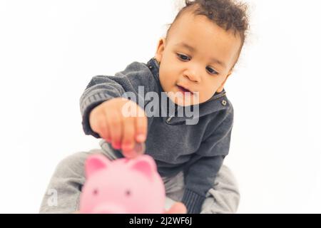 Süßes afroamerikanisches Kleinkind, das auf dem Boden sitzt und eine Münze in das rosafarbene Sparschwein-Studio-Shot mit mittlerer Vollaufnahme mit weißem Hintergrund kopiert. Hochwertige Fotos Stockfoto