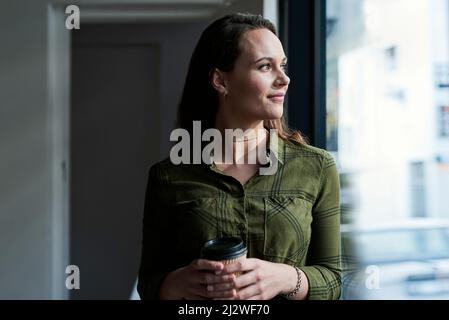Schauen Sie immer auf die hellere Seite der Dinge. Aufnahme einer attraktiven jungen Geschäftsfrau, die Kaffee trinkt und aus dem Fenster ihres Büros blickt. Stockfoto