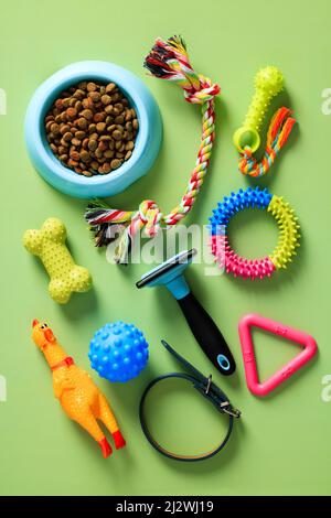 Gruppe von Haustierzubehör, Schale mit Trockenfutter, Spielzeug, Pflegemittel auf grünem Tisch. Flach liegend, Draufsicht, Overhead. Stockfoto