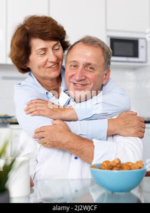 Reifes Familienpaar umarmt und sitzt am Küchentisch, während er Tee trinkt Stockfoto