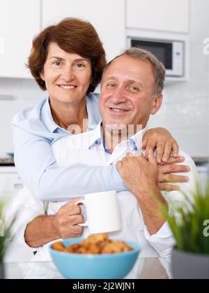 Reifes Familienpaar umarmt am Küchentisch während des Teetrinkens Stockfoto