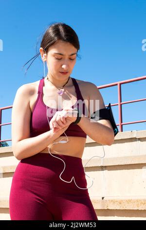 Eine junge Sportlerin, die mit einem violetten Oberteil und kastanienbraunen Leggings, einem Handy-Armband, einem Herzanhänger und Kopfhörern gekleidet ist, manipuliert ihre s Stockfoto