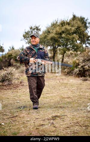 Ganzkörper gealterter männlicher Jäger in Camouflage-Oberbekleidung und Mütze, der Waffe trägt und wegschaut, während er Zeit in der herbstlichen Landschaft verbringt Stockfoto