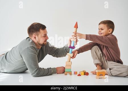 Fürsorglicher Vater hilft seinem Sohn auf weißem Hintergrund auf dem Boden zu spielen. Vater und Kind bauen Turm aus bunten Holzziegeln und haben Spaß zusammen Stockfoto