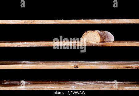 Die Hälfte des Brotlaibs wurde während der Kriegszeit in der Bäckerei auf ein leeres Holzregal mit weißem Mehl auf schwarzem Hintergrund gelegt Stockfoto