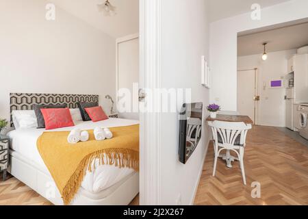 Apartment mit einem Schlafzimmer mit einem großen Bett mit Kissen und einer senffarbenen Decke neben einem Wohnzimmer mit TV und einem kleinen Holztisch Stockfoto
