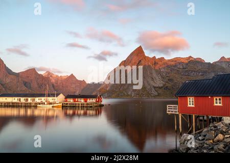 Fischerboote, rote Häuser im Hafen von Hamnoy, Moskenesoy, Lofoten, Norwegen. Sonnenlicht auf Berggipfeln. Stockfoto