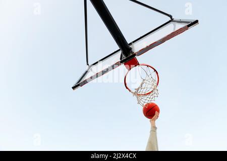 Blick von unten auf eine Hand, die einen Korb in einem Basketballkorb mit dem Himmel im Hintergrund schießt, Konzept des urbanen Sports im Freien, Platz für Text kopieren Stockfoto