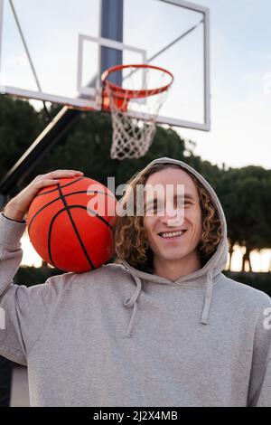 Vertikales Porträt eines lächelnden jungen Mannes, der auf einem Stadtgericht einen Basketball hält, Konzept des urbanen Sports auf der Straße, Textraum kopieren Stockfoto