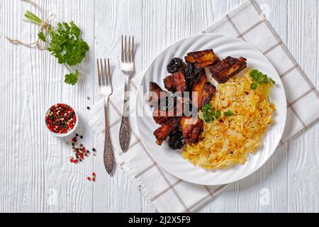 Geschmortes Sauerkraut mit Schweinefleisch und Pflaumen auf weißem Teller auf Holztisch, horizontale Ansicht von oben, flaches Lay, Freiraum Stockfoto