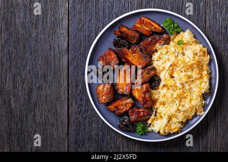 Nahaufnahme von geschmortem Sauerkraut mit Schweinebraten, kurzen Rippen und Pflaumen auf Teller auf dunklem Holztisch, horizontale Ansicht von oben, flaches Lay, freier Platz Stockfoto