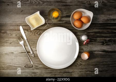 Eine Draufsicht auf drei rote Eier in Becher, Messer, Gabel, einer Zuckerplatte und einem zerbrochenen Eierjoch in einem durchsichtigen Glas auf einem grauen Holzküchentisch Stockfoto