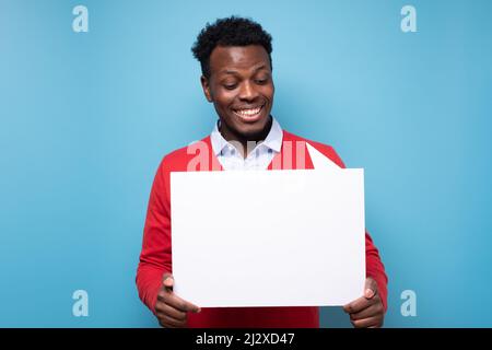 Junger lächelnder afrikaner, der weiße, leere Tafel an der blauen Wand hält. Stockfoto
