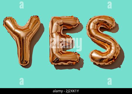 Einige goldene, buchstabenförmige Luftballons bilden das Wort „Ja“ auf blauem Hintergrund Stockfoto