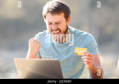 Aufgeregt Mann hält Kreditkarte online kaufen auf Laptop feiert Erfolg im Freien Stockfoto