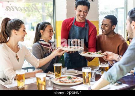 Vertrauter Kellner serviert leckere Pizza margherita an multikulturelle Freunde in gemütlichen Pizzeria Restaurant - multiethnische Freunde, die Spaß zusammen haben Stockfoto