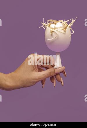 Ein einfaches Konzept von Ostern. Die Hälfte einer weißen Eierschale ist mit hellem Heu verziert. Eine weibliche Hand hält ein weißes Ei auf einem Eisstock. Der Hipster ide Stockfoto