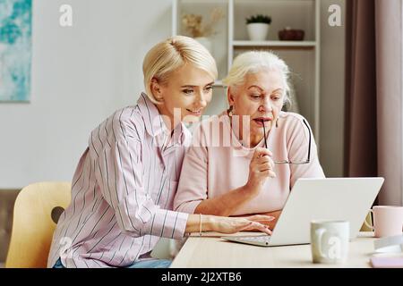 Porträt einer blonden jungen Frau, die ältere Mutter mit Laptop und Internet in einem gemütlichen Zuhause unterrichtet Stockfoto
