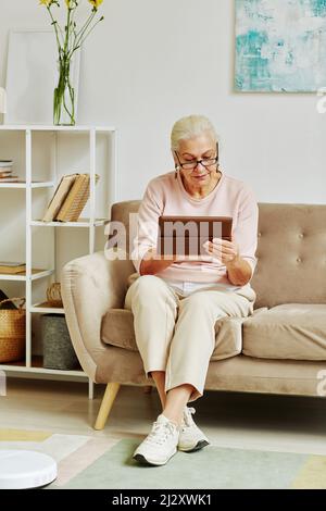 Vertikale Ganzkörperporträt der modernen älteren Frau mit Tablet, während sie auf dem Sofa im weißen Innenraum sitzt Stockfoto