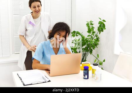 Porträt von zwei schönen jungen weiblichen Mixed-Race-Doktoren am Laptop, während sie im Krankenhaus lächeln und Spaß haben Stockfoto