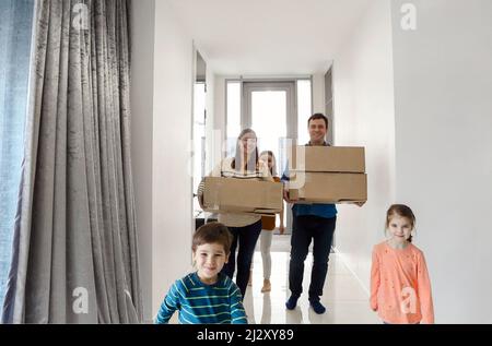 Glückliche Familie, die in der Halle läuft. Lustige Kinder springen mit Karton-Box, Eltern lachen, spielen mit Kindern Spaß haben zusammen bewegen sich im neuen Haus. Hypothek Stockfoto