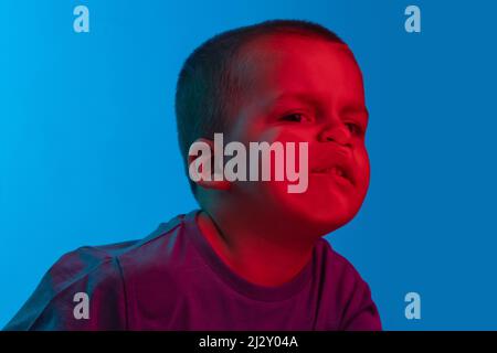 Nahaufnahme kleiner Junge, niedliches Kind lehnt an transparentem Glas durch Wange isoliert auf blauem Studiohintergrund in Neonlicht Stockfoto