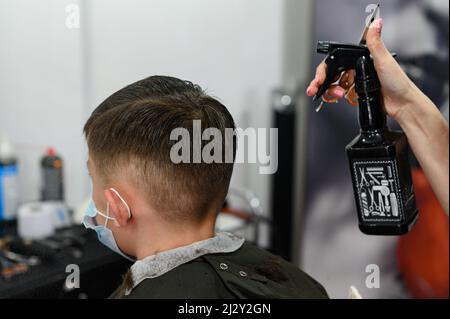 Ein Teenager in einem Schönheitssalon bekommt einen Haarschnitt, ein Friseur schneidet einem Teenager die Haare und benetzt seine Haare mit Spray. Stockfoto