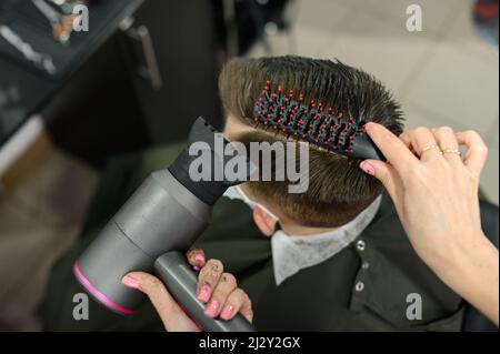 Teenager bekommt einen Haarschnitt während einer Pandemie in einem Friseurladen, einen Haarschnitt und trocknet Haare nach einem Haarschnitt, styling Haare nach einem Haarschnitt mit einem Haartrockner. Stockfoto